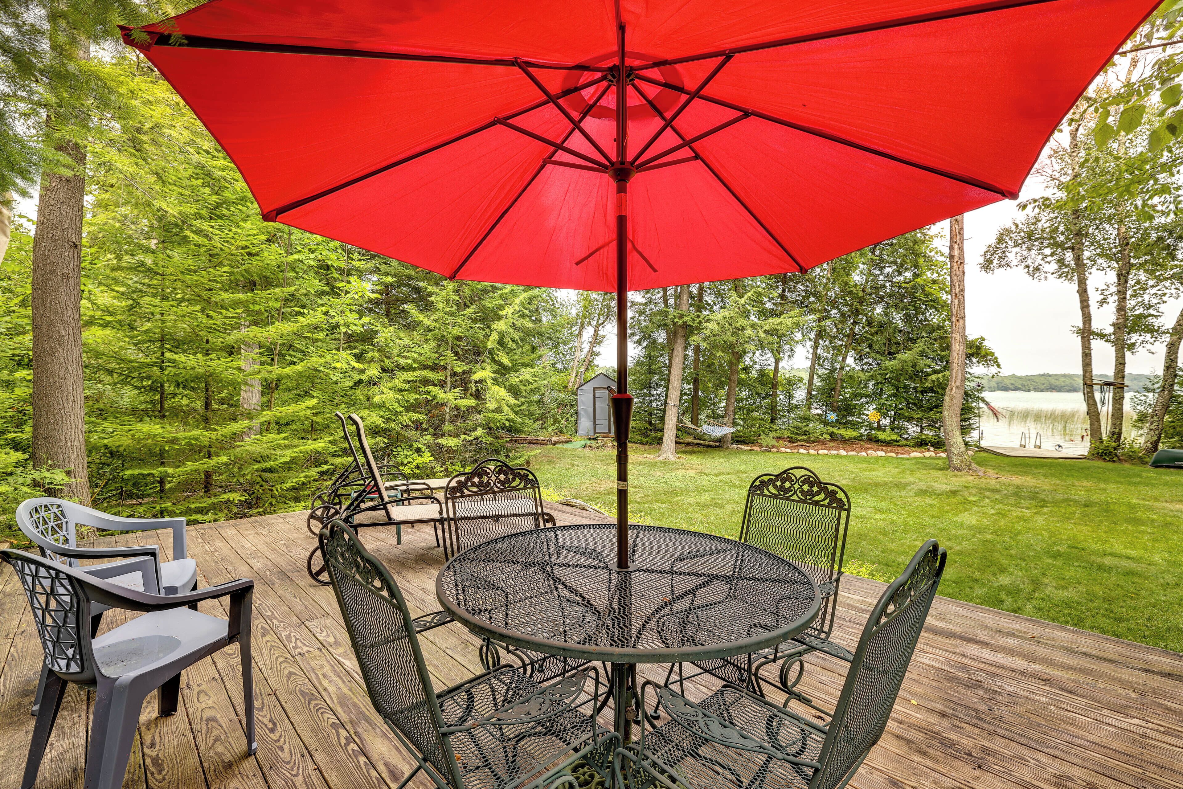 An outdoor patio with a round metal table and chairs, a red umbrella, and a view of a lake surrounded by trees.