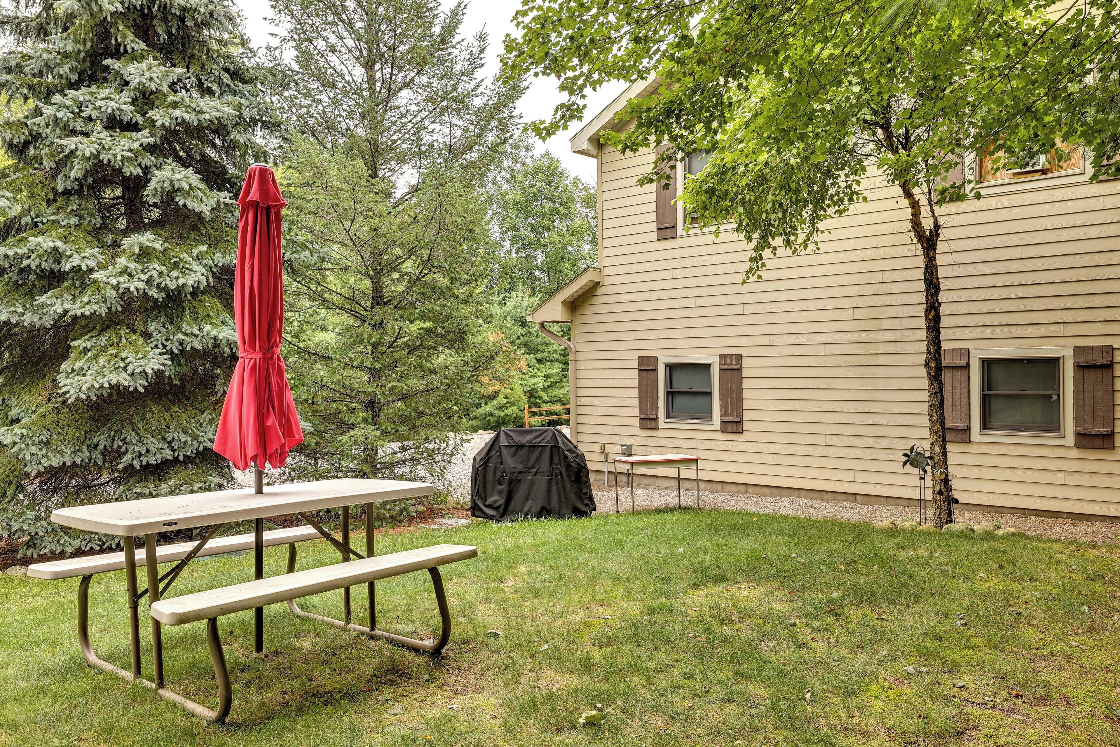 A picnic table with a red umbrella, a grill, and a house with a tree in the yard.
