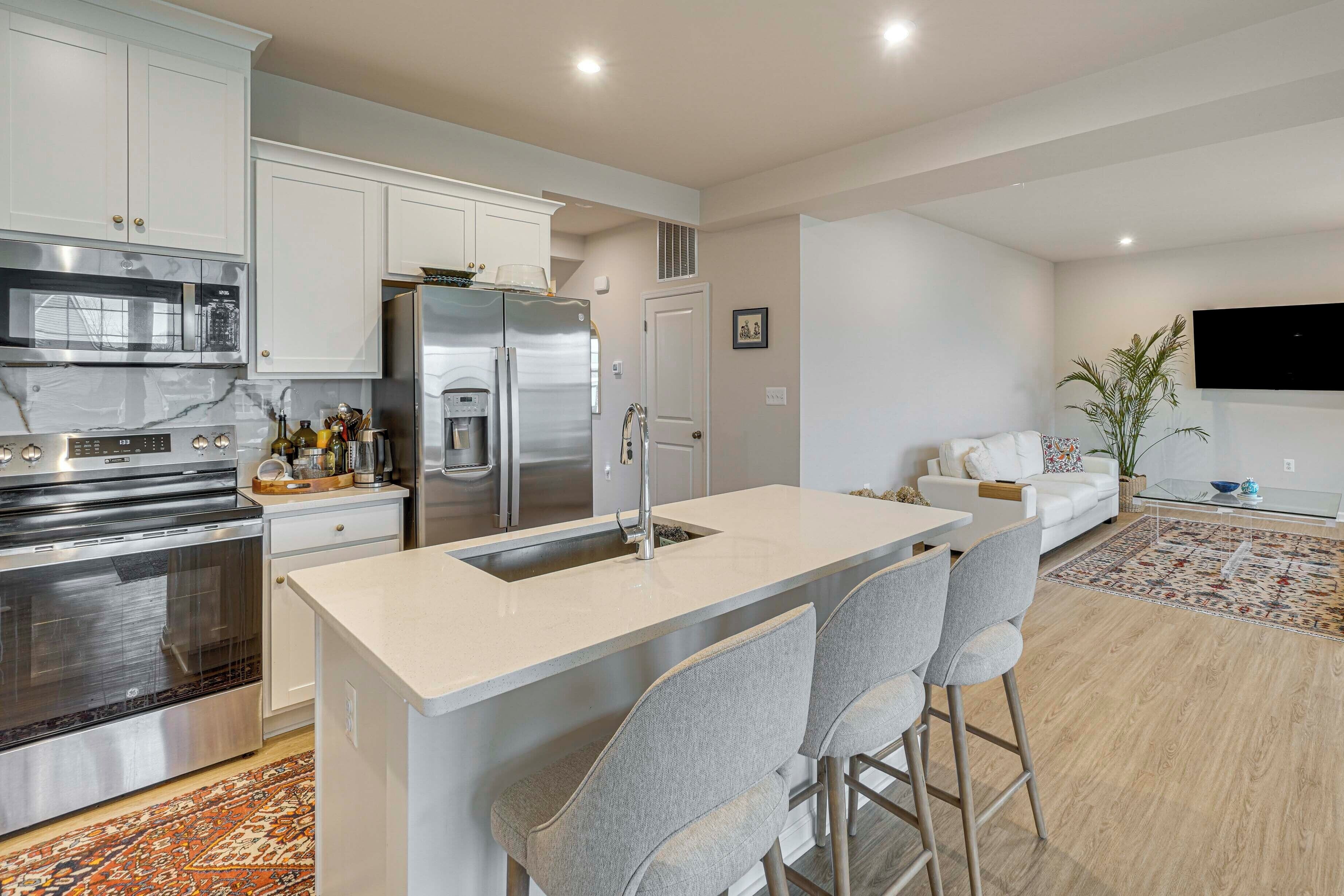 A modern kitchen with stainless steel appliances, a central island with bar stools, a dining area with a sofa, and a flat-screen TV mounted on the wall.