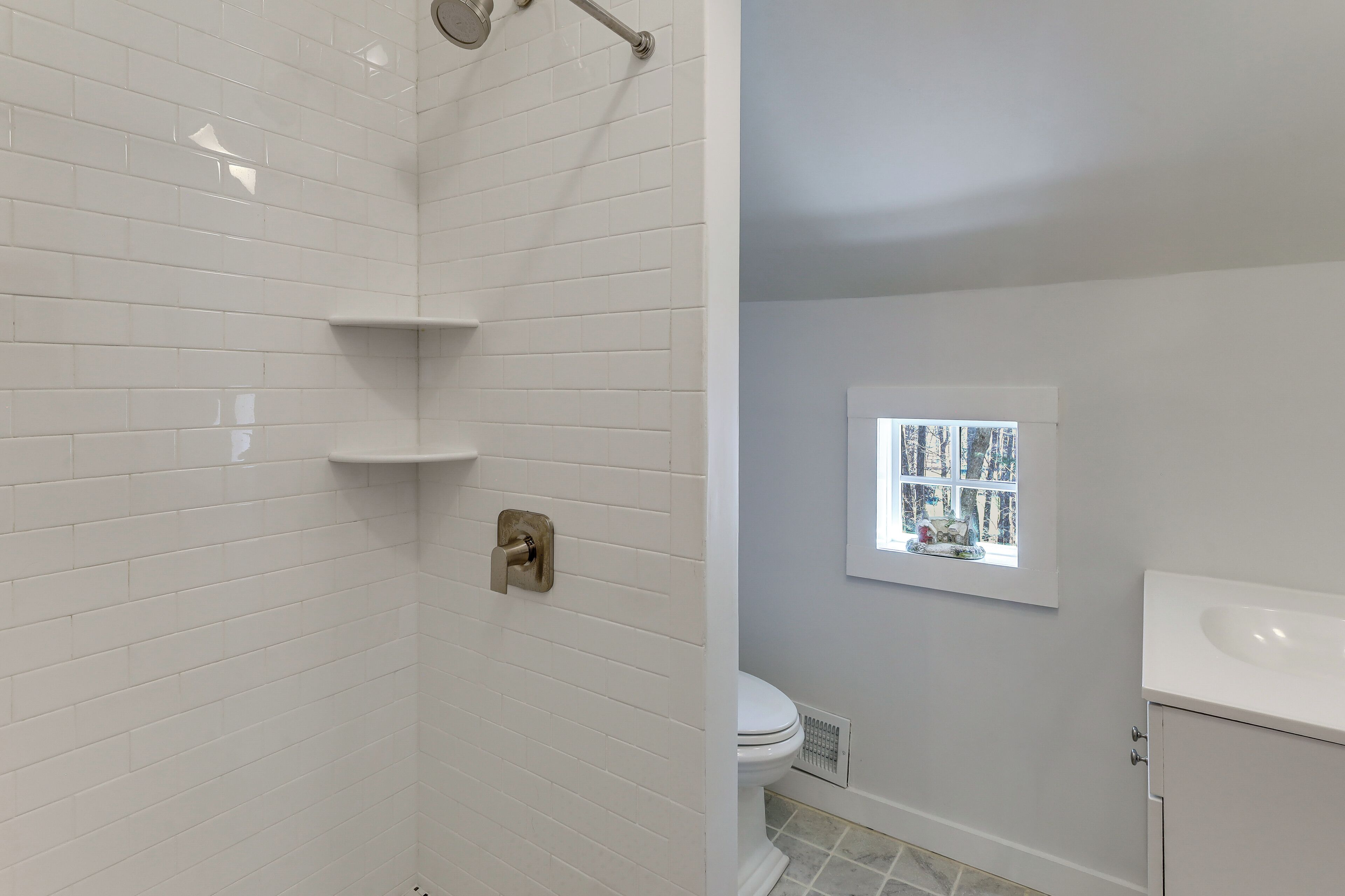 Une salle de bain avec des carreaux de céramique blancs rectangulaires, une douche, des toilettes, une fenêtre avec vue et un lavabo.