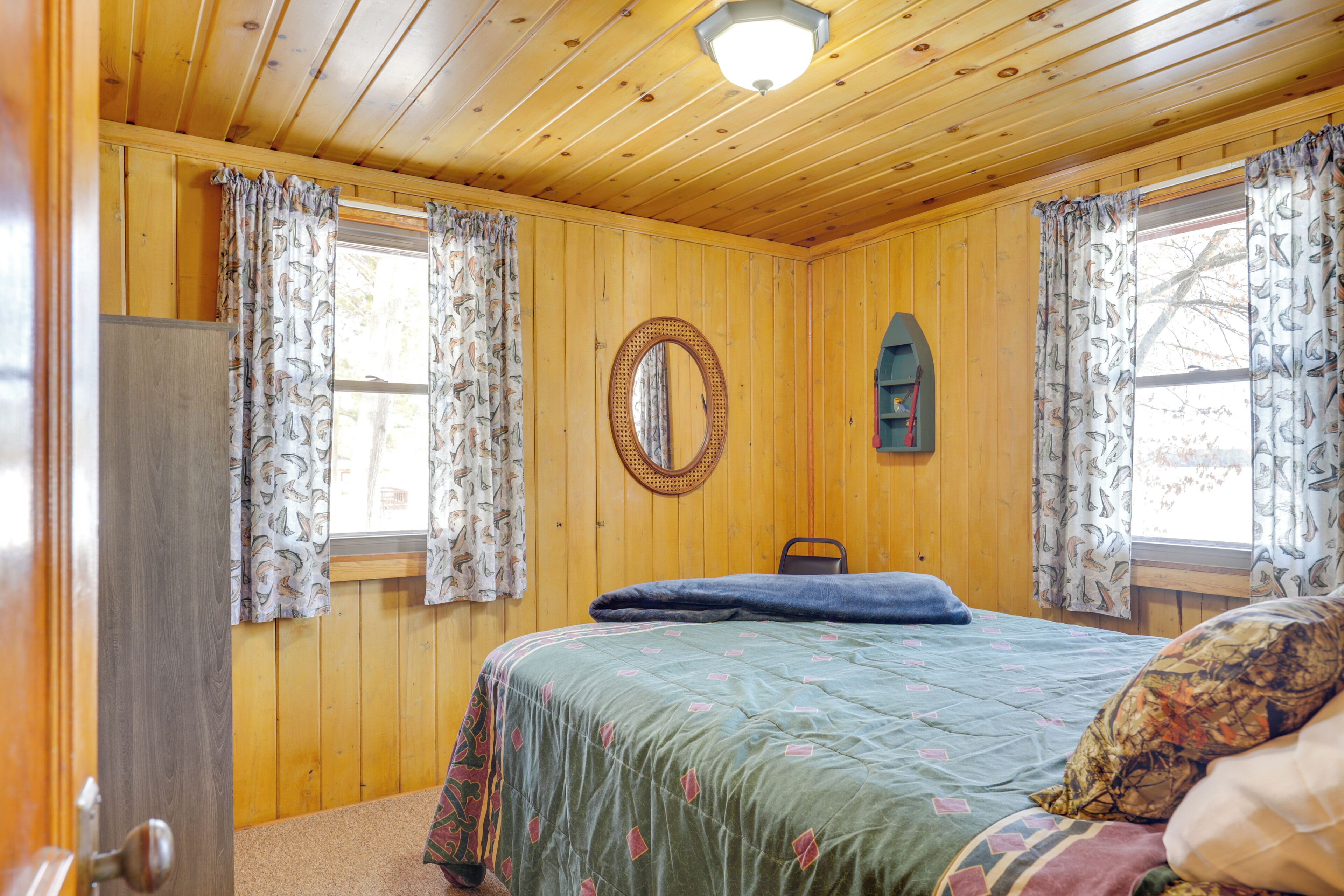A room with wooden walls, a bed with a patterned bedspread, a round mirror, and curtains with a fish pattern.