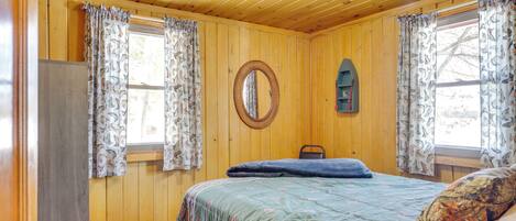 A room with wooden walls, a bed with a patterned bedspread, a round mirror, and curtains with a fish pattern.