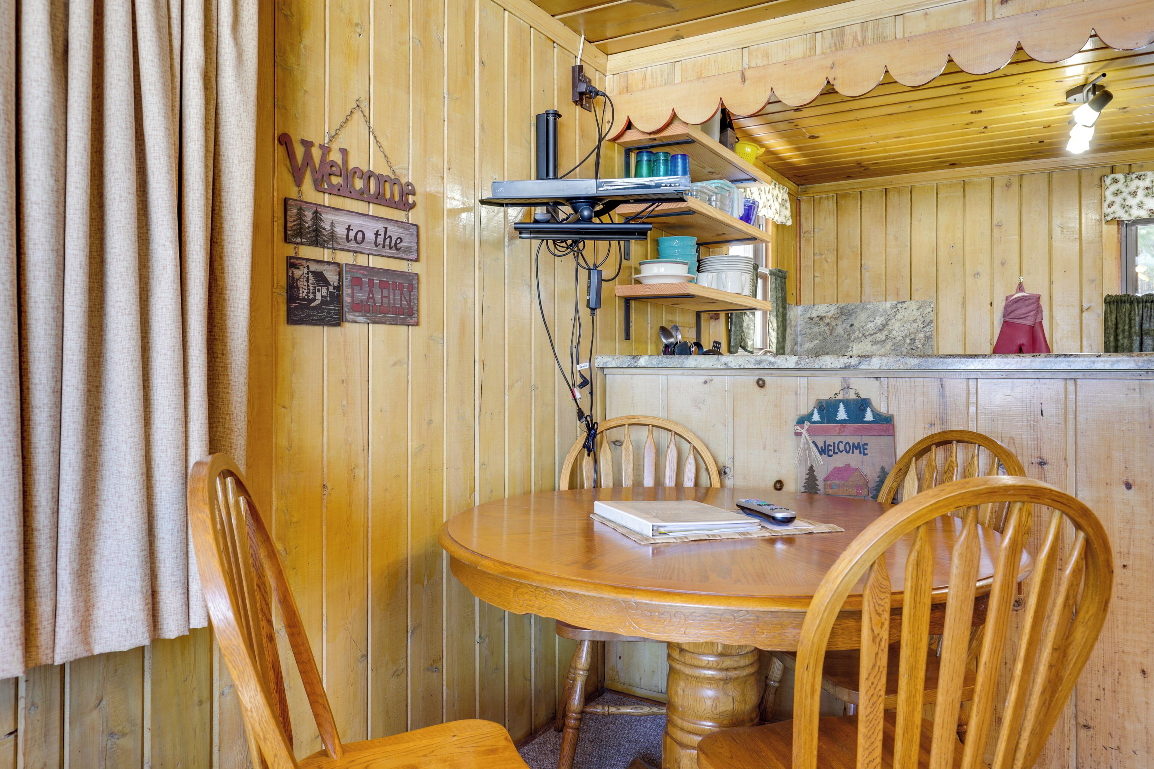 A wooden interior with a round table, chairs, a shelf with dishes, and a welcome sign.