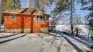 En liten, envåningsstuga med ett sluttande tak, ett fönster och en dörr, omgiven av snö och träd.
