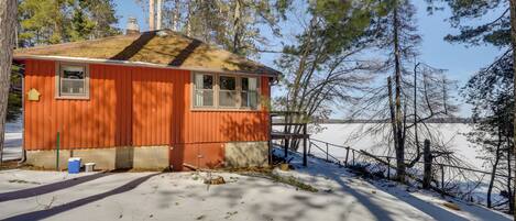 A small, single-story cabin with a sloped roof, a window, and a door, surrounded by snow and trees.