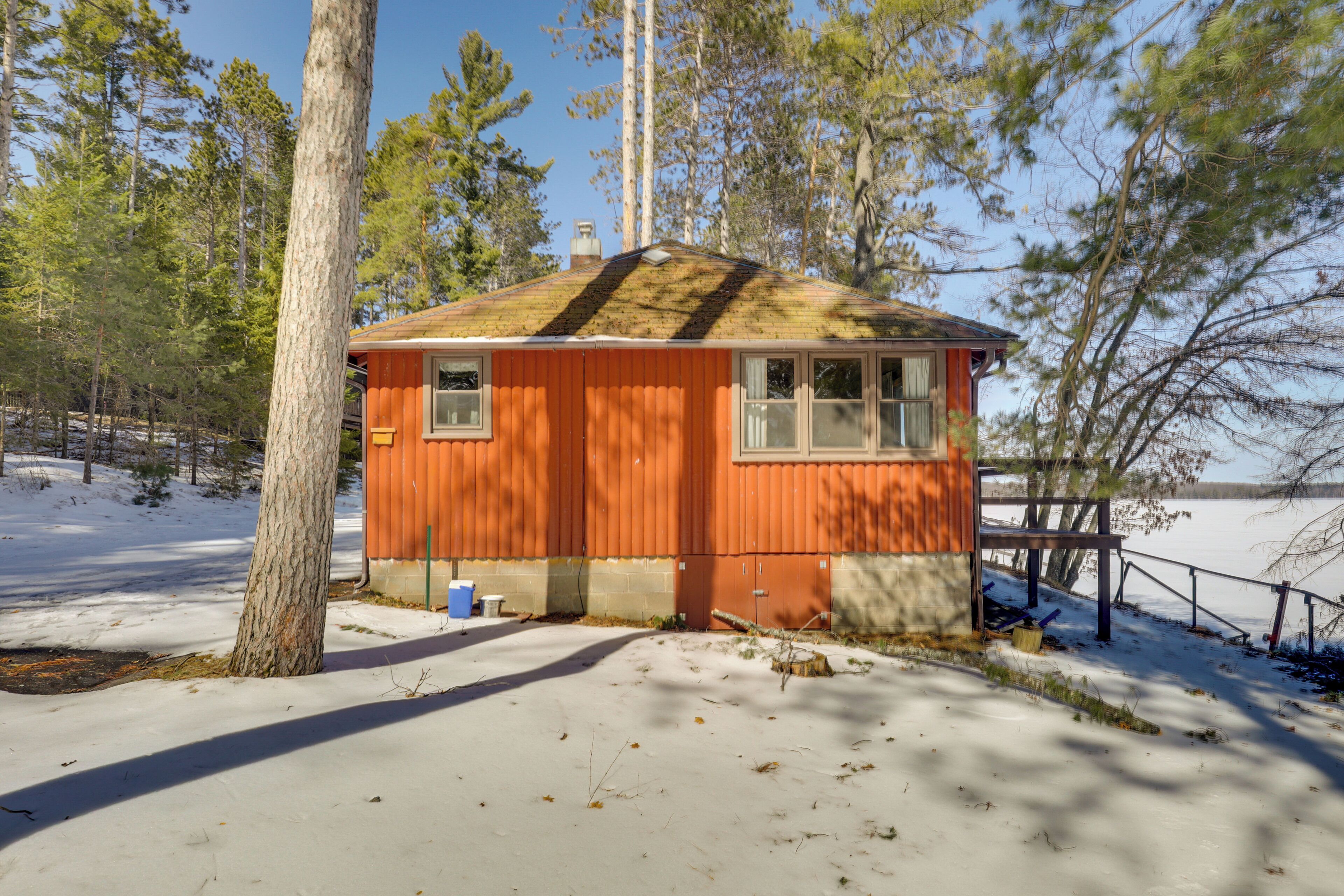 A small, single-story cabin with a sloped roof, surrounded by trees and snow, near a body of water.