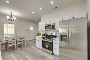 Una cocina moderna con armarios blancos, electrodomésticos de acero inoxidable, una zona de comedor con mesa y sillas de madera, y una araña.