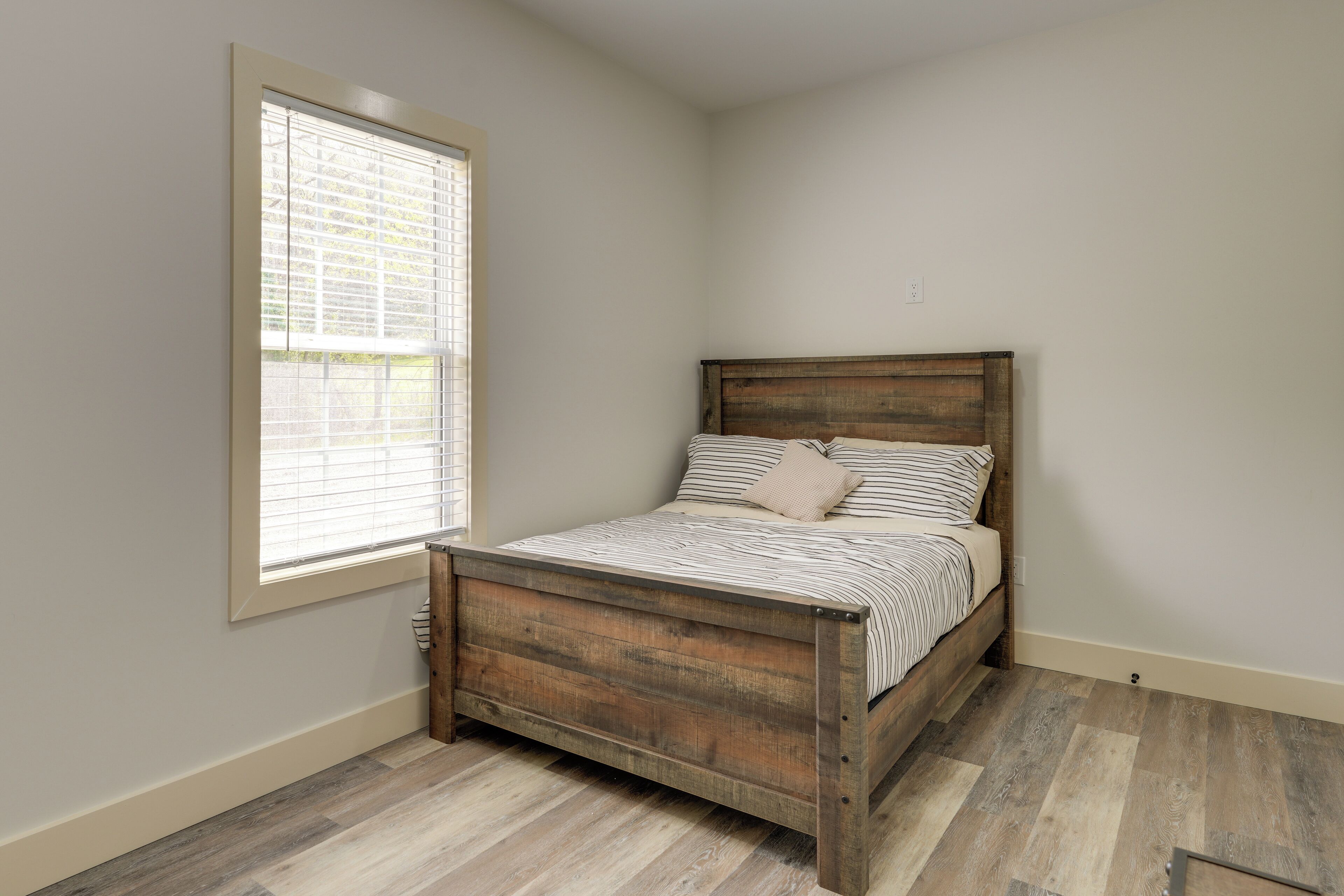 Una cama individual con estructura de madera y ropa de cama a rayas en una habitación con ventana y suelo de madera.