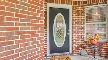 A brick house with a dark door, a welcome sign, a window with blinds, and a potted plant on a small table.