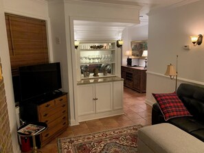 Un espace de vie compact comprenant un téléviseur, une petite table avec des magazines, un canapé agrémenté d’un plaid, et un coin cuisine avec des placards et un plan de travail.
