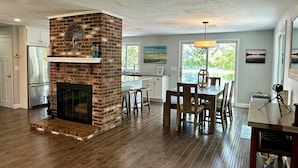 A spacious living area with a brick fireplace, wooden flooring, a dining table with chairs, and a kitchen with white cabinets. - Located directly on Pond, Swimming, Kayaking, Fishing, Beach Pass, A/C, Clean! (Barnstable)
