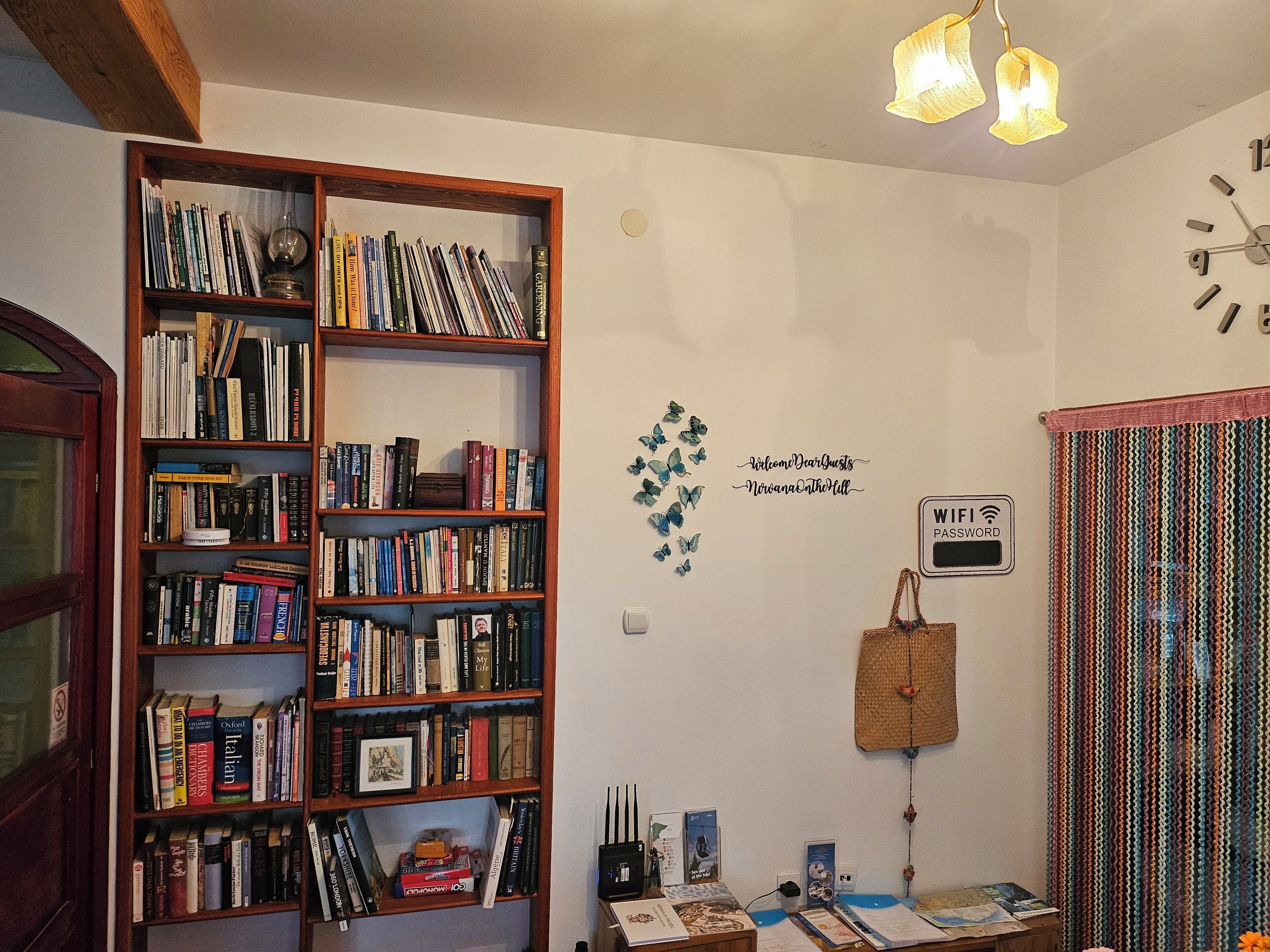 A room with a bookshelf filled with books, a wall with a welcome sign, a WiFi password sign, and a patterned curtain.