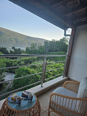 Ein Balkon mit Blick auf einen See und Hügel, mit Korbmöbeln und einem Tisch mit Tasse und Gebäck.