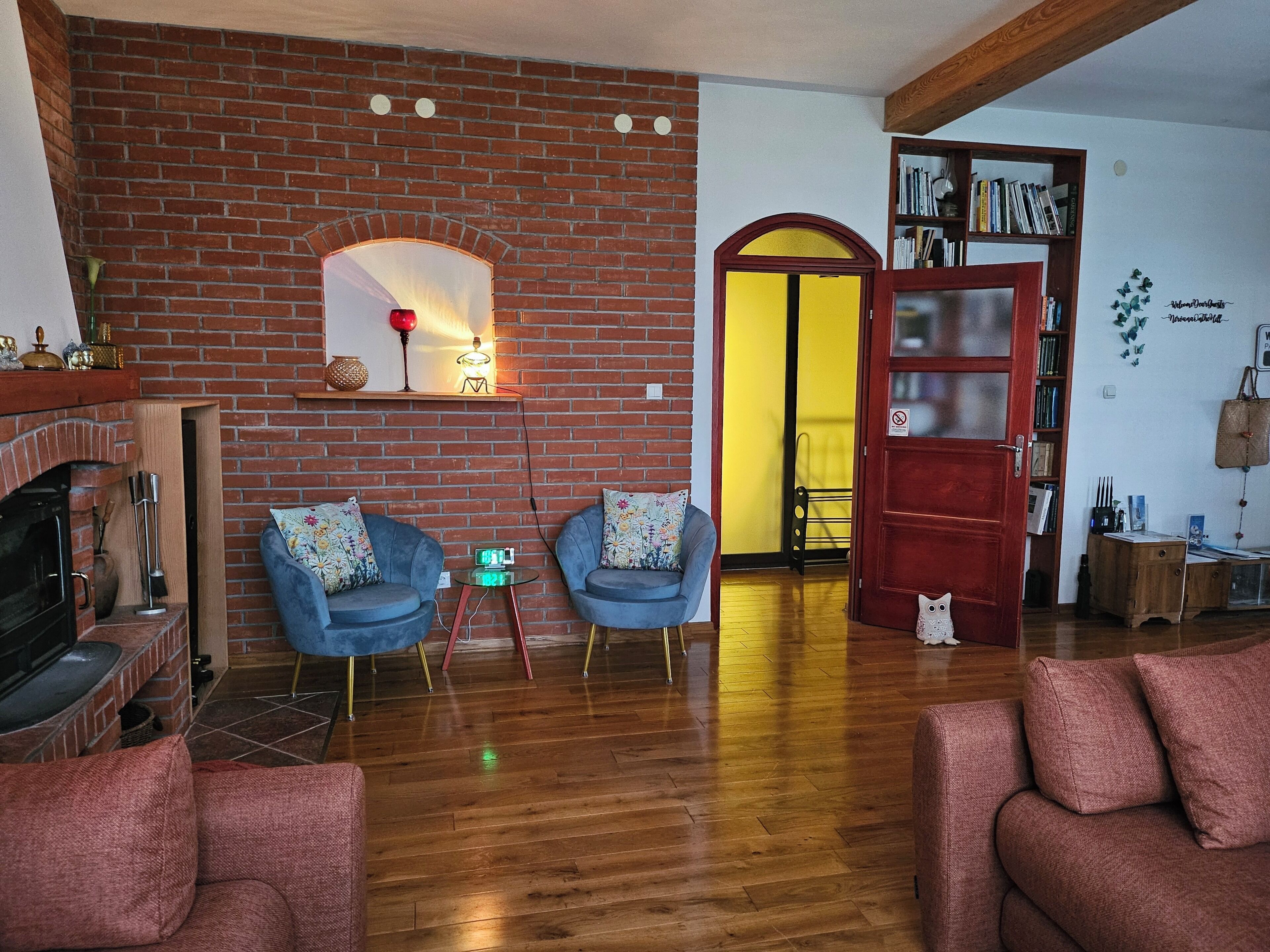 Una sala de estar con chimenea de ladrillo, dos sillones azules, un sofá rojo, piso de madera y una estantería.