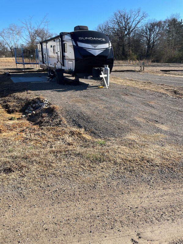 A black and white RV with 'SUNDANCE' written on the back, parked on a gravel surface with dry grass and trees in the background.