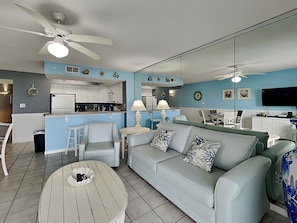 A modern living room with a sofa, chairs, a coffee table, a TV, and a kitchen in the background. - Gulf Dunes #206 East | Resort Amenities + Walkable to Dining/Activities (Fort Walton Beach)