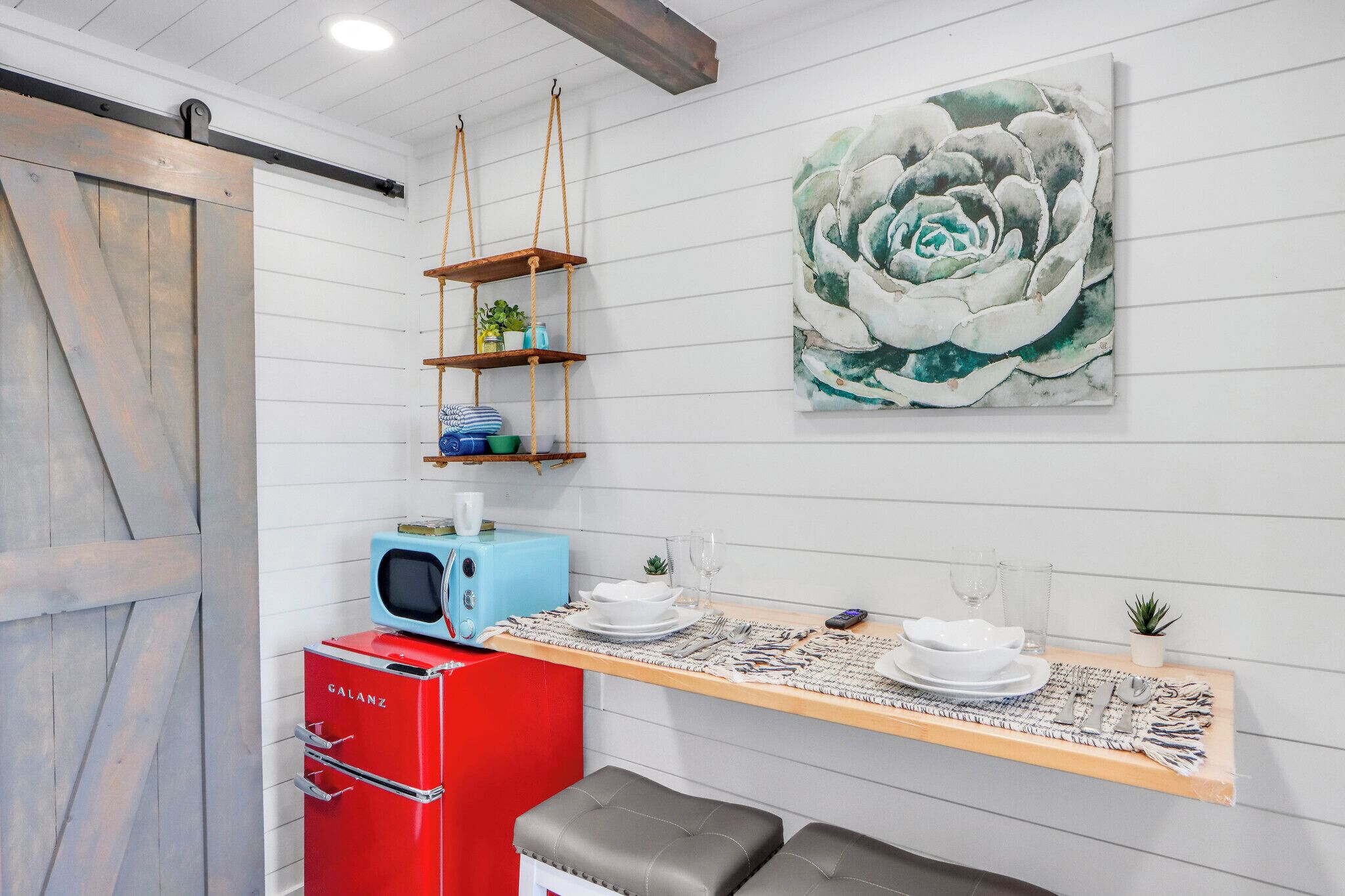Een keuken met een rode Galanz koelkast, een blauwe magnetron, een houten tafel met een placemat en een groot bloemenkunstwerk aan de muur.