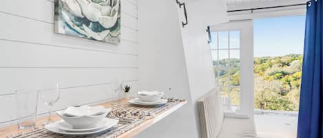 Un comedor con una mesa preparada para dos, vistas al exterior y un cuadro grande con motivos florales en la pared.