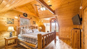 Ein Schlafzimmer in einer Blockhütte mit einem hölzernen Bett, zwei Nachttischen mit Lampen, einem an der Wand montierten Fernseher und einem Deckenventilator.