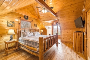 A log cabin bedroom with a wooden bed, two bedside tables with lamps, a television mounted on the wall, and a ceiling fan. - 4BR, Views, Firepits, Hot Tub! (Pigeon Forge)