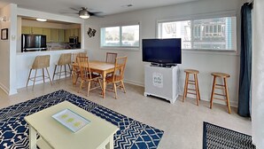 Sala de estar con televisor, mesa de comedor, sillas, una kitchenette pequeña y un tapete estampado.
