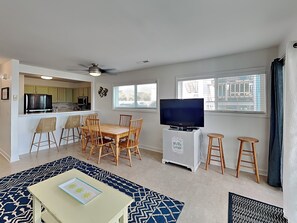A living room with a TV, dining table, chairs, a small kitchenette, and a patterned rug. - The Riggings #E1 | Pool + Beach Access (Kure Beach)