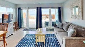 Una sala de estar con sofá, televisor, mesa y vistas a la playa a través de amplios ventanales correderas.