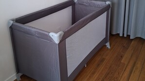 Un parc pour bébé d’angle, doté d’une paroi en tissu ajouré gris, placé contre un mur blanc et un plancher en bois, avec un rideau gris partiellement visible.