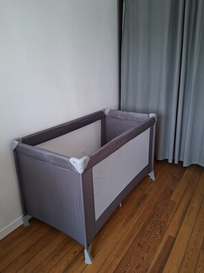 A corner baby playpen with a grey mesh side, positioned against a white wall and wooden floor, with a grey curtain partially visible. - Cosy house for 2-3 people, garden, free parking, in the heart of the village and close to hiking trails! (Signy-l'Abbaye)