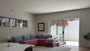 A modern living room with a sectional sofa, a dining table with chairs, and a sliding glass door leading to a patio with plants. - Bheka Guest House Parktown North, Johannesburg (Johannesburg)