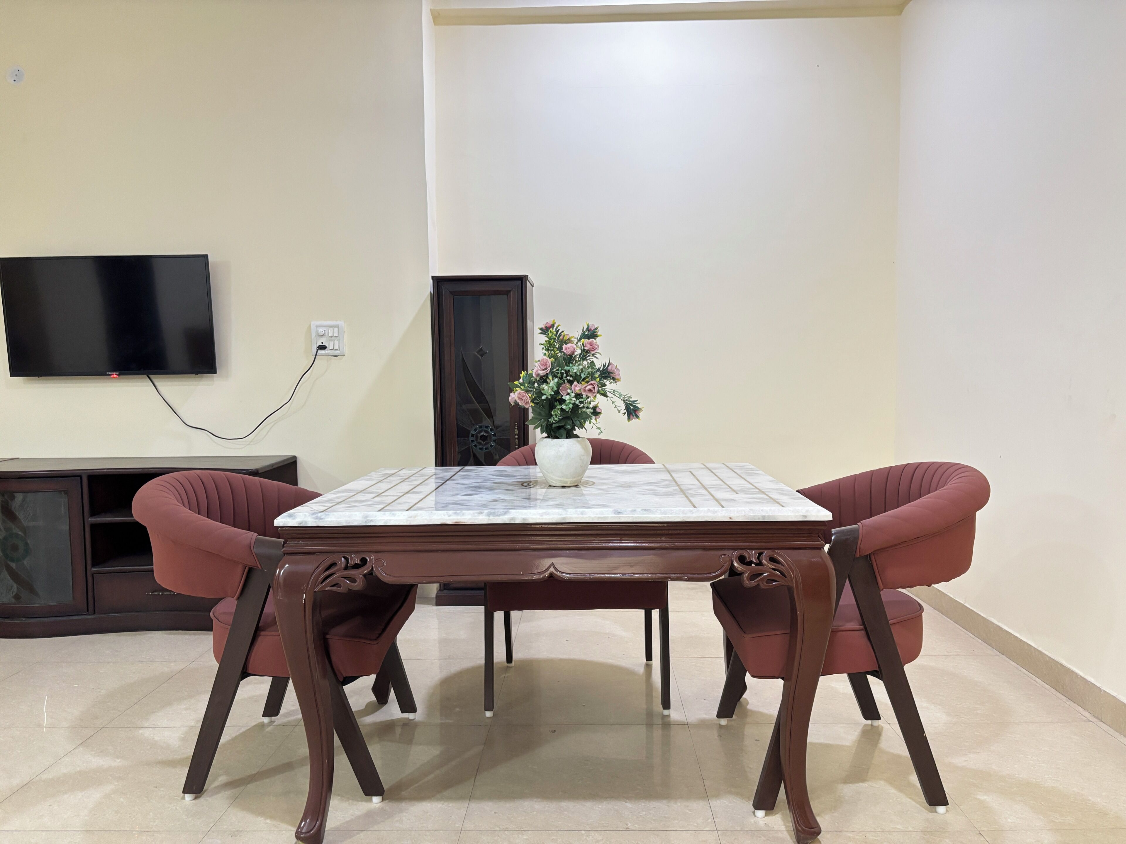 A dining area with a marble table, two chairs, a television mounted on the wall, and a flower vase.