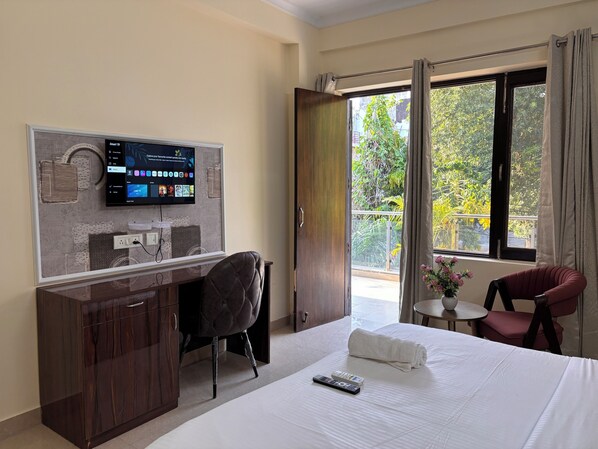 Habitación de hotel con cama, escritorio con silla, televisor montado en la pared y balcón con vistas a zonas verdes.