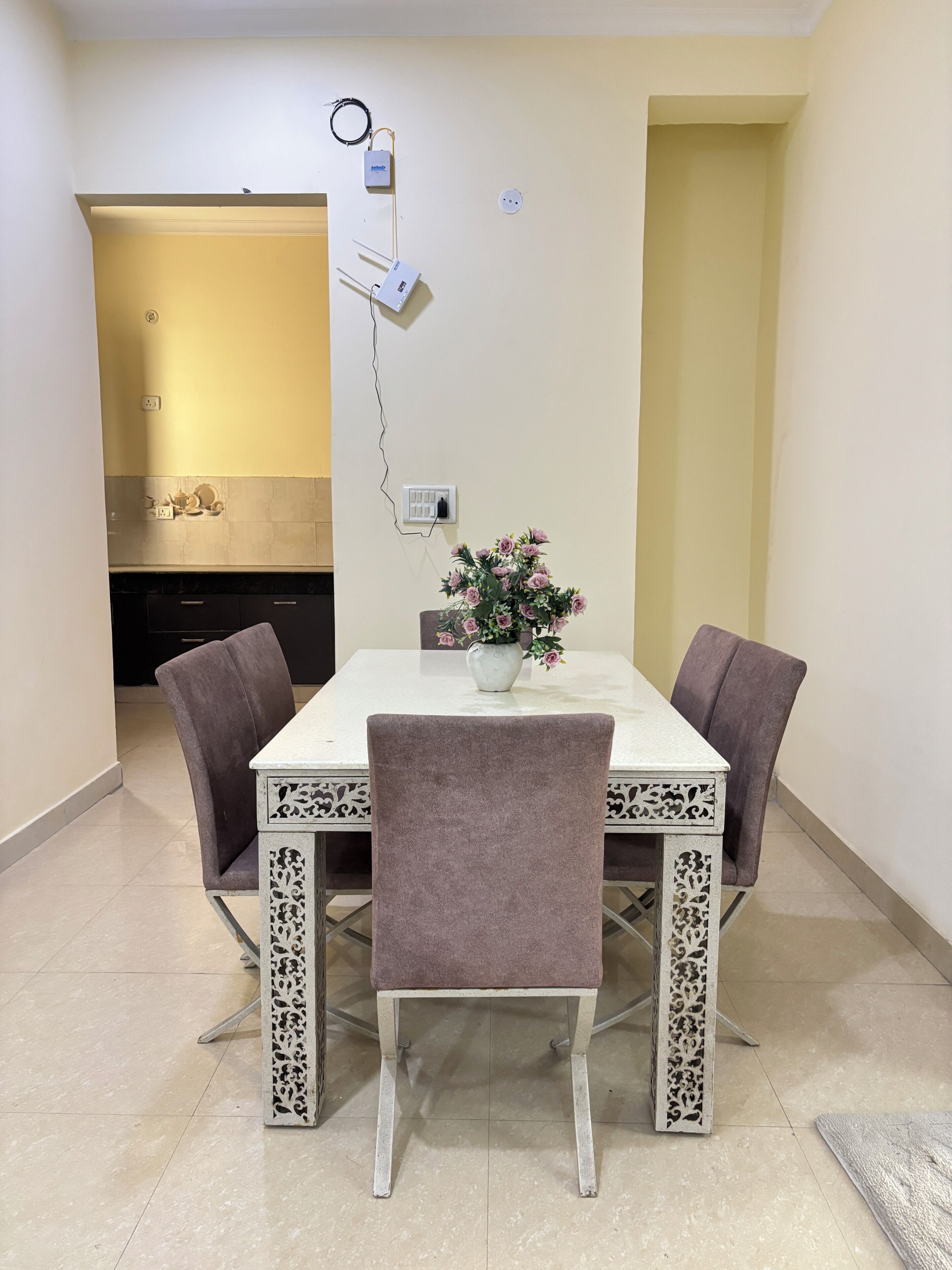 A dining area with a white table and four chairs, a vase with flowers, and a kitchen in the background.