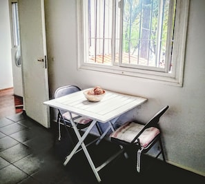 Un petit coin repas avec une table et deux chaises, un bol de nourriture, une fenêtre avec des barreaux et une porte dotée d’un éventail.