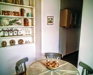 Un coin repas avec une table ronde, des chaises et un saladier rempli de nourriture. On y trouve des étagères avec des objets décoratifs et un tableau encadré accroché au mur.