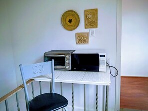 Une petite cuisine équipée d’un grille-pain et d’un four à micro-ondes, le tout posé sur un plan de travail blanc, une chaise à ossature métallique et des assiettes décoratives accrochées au mur.