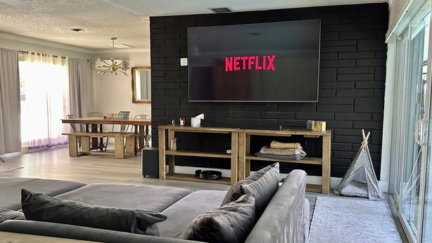 Un salon avec un mur en briques noires, un grand téléviseur à écran plat, un canapé gris, des meubles en bois et une fenêtre donnant sur un paysage agréable.