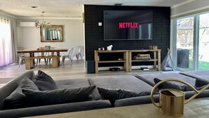 Un salon doté d’un grand téléviseur à écran plat fixé sur un mur en briques noires, d’une table basse en bois, d’un canapé gris et d’un coin repas avec une table et des chaises en bois.