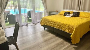 Une chambre à coucher comprenant un lit, une chaise, une table de chevet et donnant sur une piscine grâce à une porte vitrée.