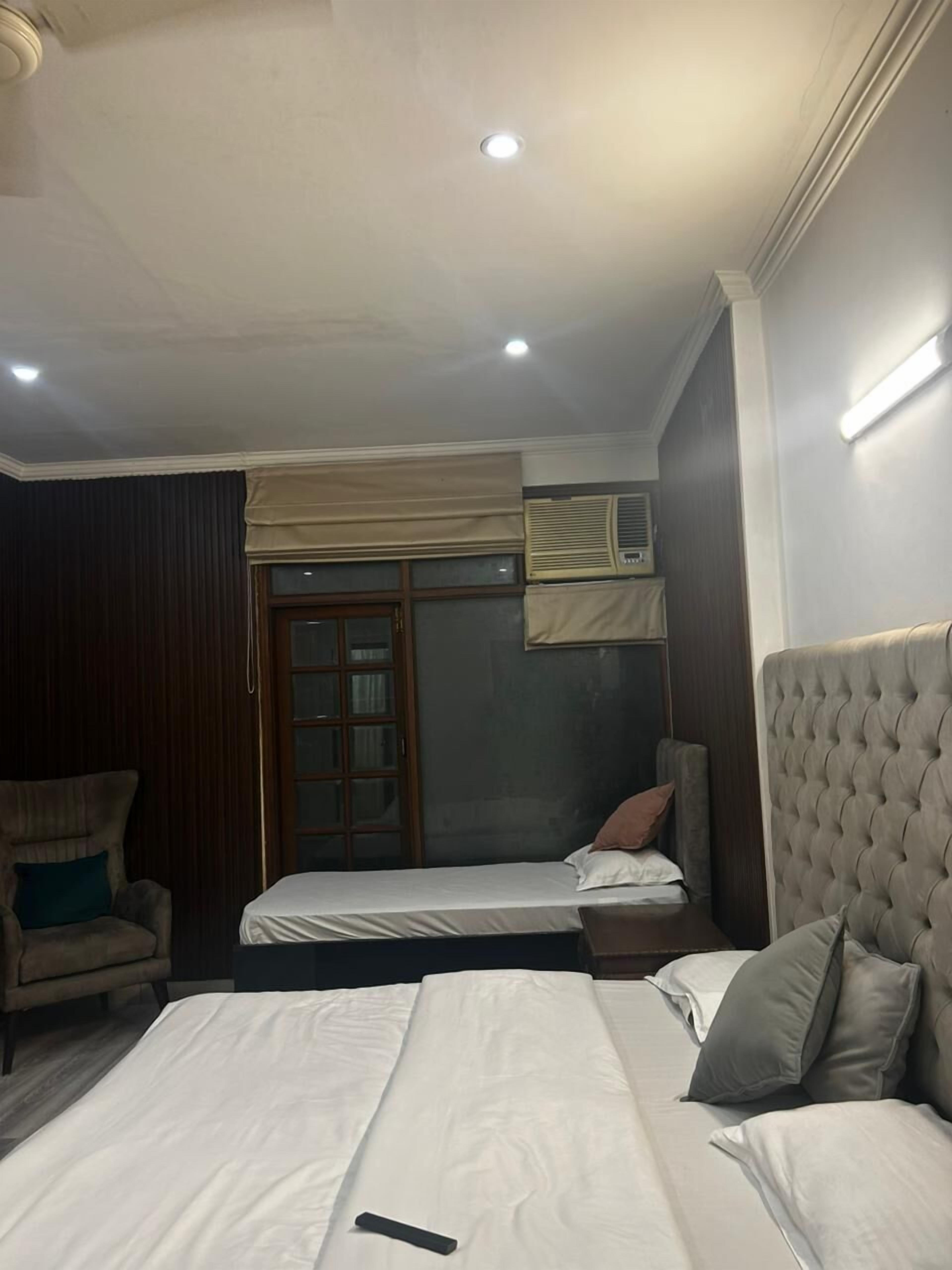 A hotel room with two beds, a chair, a window with curtains, an air conditioning unit, and a ceiling light.
