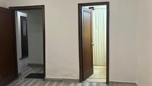 Een kamer met een houten vloer, een deur die naar een andere kamer leidt, en een zichtbare lichtschakelaar aan de muur.