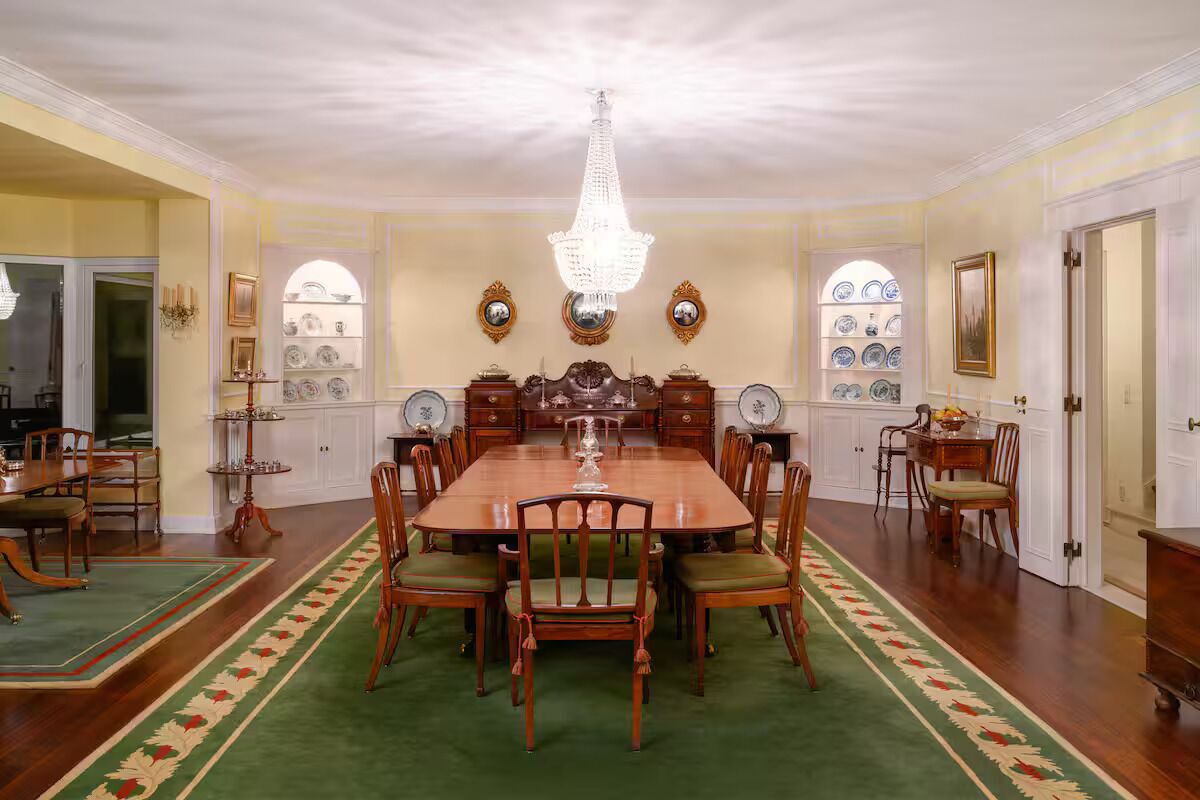 A formal dining room with a large wooden table, green upholstered chairs, a chandelier, and decorative wall shelves.