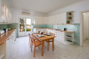A modern kitchen with white cabinets, a dining table set for four, and a view through the glass door. - Villa in Quinta do Perú Golf & Country Club (Quinta do Conde)