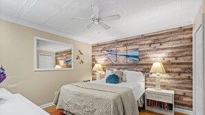 Une chambre avec un mur en bois, un ventilateur de plafond, un lit recouvert d’une courtepointe, deux tables de chevet avec des lampes et une bibliothèque.