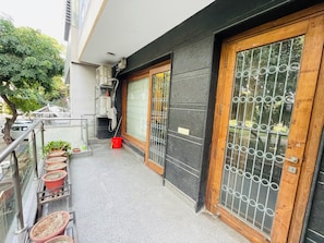 A balcony with potted plants, a red bucket, and a black door with a metal gate. - Fortune Home Service Apartment 4BHK J191 (New Delhi)