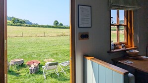 Een open deur naar een kamer met uitzicht op een grasveld, een raam met uitzicht, een kleine tafel met stoelen, en een wandlamp.