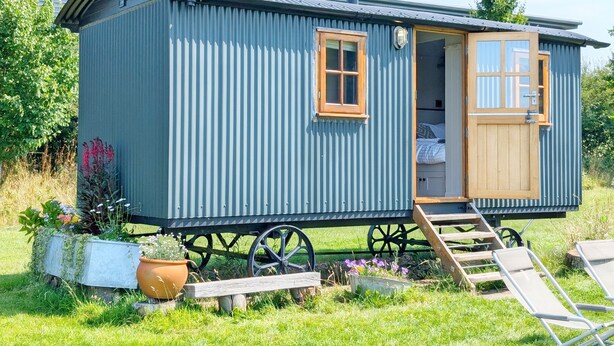 Een klein, blauw, gelijkvloers huis met een houten deur en ramen, een schoorsteen, en een tuin met planten en een bankje.