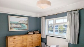 Une chambre à coucher avec une commode en bois, un tableau représentant un paysage côtier, une fenêtre avec des rideaux et un lit avec des serviettes.