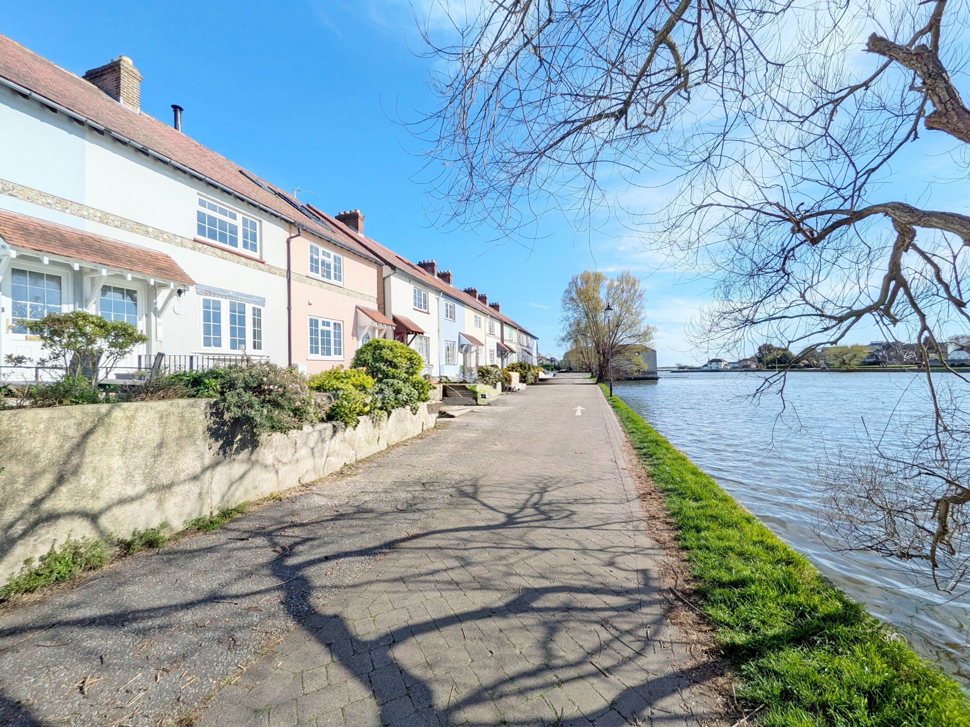 A paved walkway alongside a river, with houses on one side and a grassy embankment on the other.