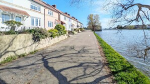 Un chemin pavé longe une rivière, avec des maisons d’un côté et une berge herbeuse de l’autre.